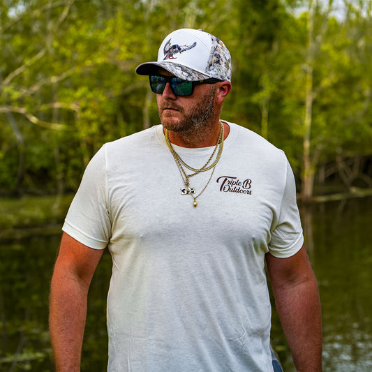 Zach Rushing wearing a white crewneck t-shirt with a 'Triple B Outdoors' logo on the chest, a white camouflage baseball cap, and sunglasses, standing outdoors in front of a body of water with green trees.