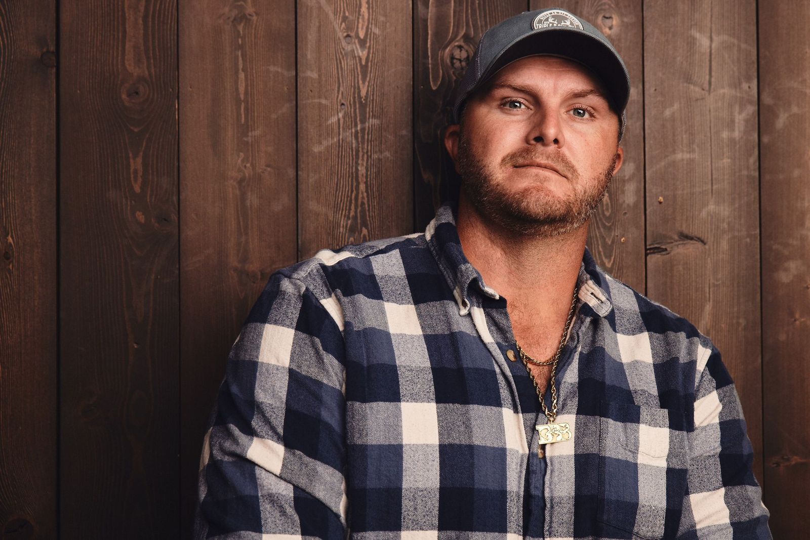 Zach Rushing smirking while leaning against a wood panel wall while wearing flannel and a bbb outdoors hat and necklace.